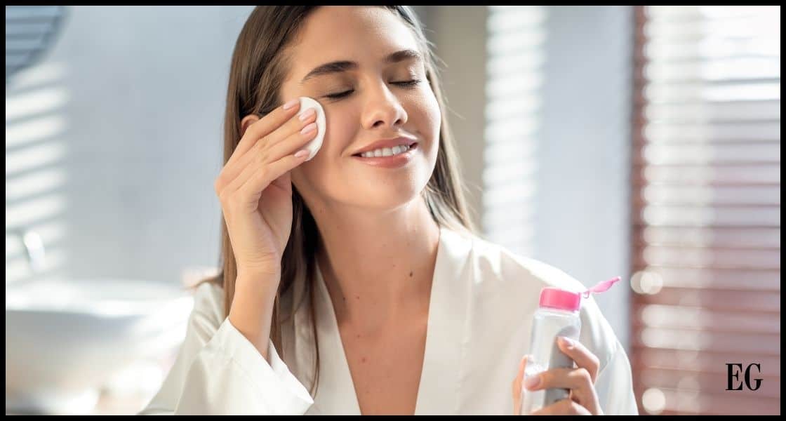 Imagen de una mujer sonriente aplicando tónico facial o removedor de maquillaje en su rostro con un algodón. Ella tiene los ojos cerrados y sostiene una botella con dispensador en la otra mano. La mujer viste una bata o camisa blanca y parece estar en un entorno interior bien iluminado, posiblemente su hogar.