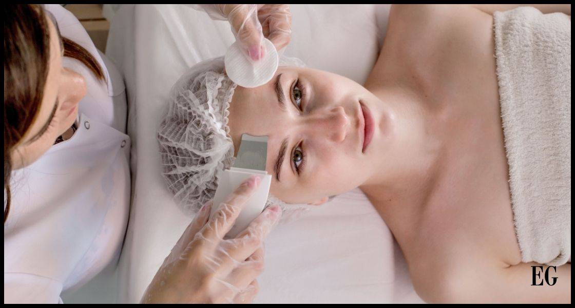 Una mujer acostada, recibiendo un tratamiento facial por parte de otra persona que se inclina sobre ella. La mujer tumbada lleva una banda protectora en el cabello y tiene una expresión relajada en su rostro. La persona que realiza el tratamiento lleva guantes y está utilizando un aplicador o dispositivo en la frente de la mujer acostada. Ambas parecen estar en un entorno profesional, posiblemente un salón de belleza o clínica de cuidado de la piel.